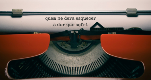 Foto de uma máquina de escrever com um papel contendo a frase "quem me dera esquecer a dor que sofri".