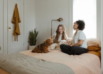 Nem toda zona de conforto é ruim. Foto de duas mulheres sentadas sobre uma cama acompanhadas de um cachorrinho.