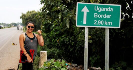 Jéssica Paula apontando para uma placa onde está escrito "Fronteira de Uganda" a 2,9 km