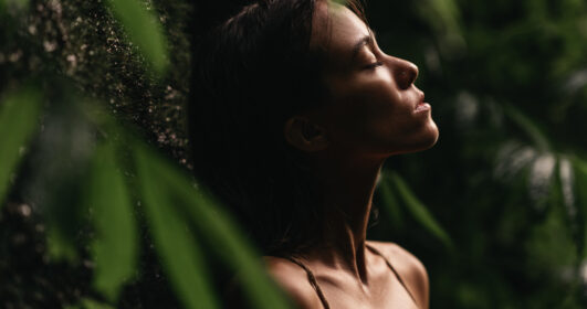 Retrato de uma jovem próxima a plantas com os olhos fechados.