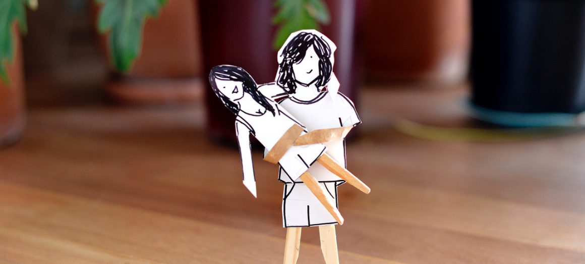Na imagem, dois bonequinhos de madeira e papel desenhados pela colunista Estéfi. Um deles, de pé, representando seu filho, está segurando a mãe no colo.