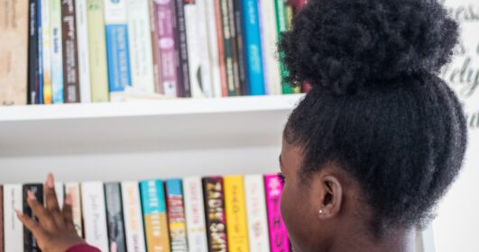 Foto de uma jovem de costas em frente a uma estante repleta de livros.