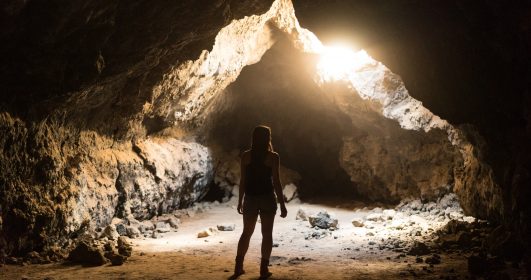 Imagem mostra uma mulher parada em frente a entrada de uma caverna escura,