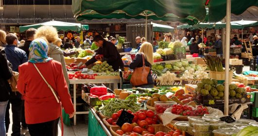Feira livre com diversas frutas, legumes e verduras e pessoas fazendo compras.