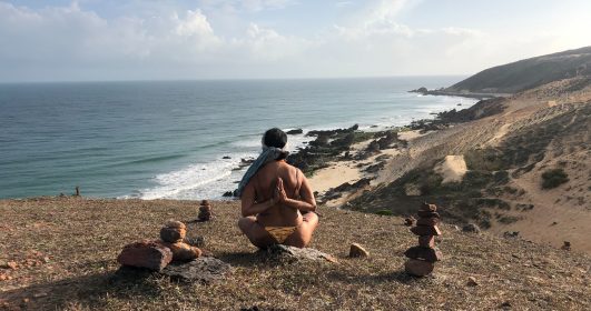 Pessoa praticando meditação na praia, céu limpo, claro e mar azul com ondas baixas. Técnicas de respiração para ajudar a meditar.