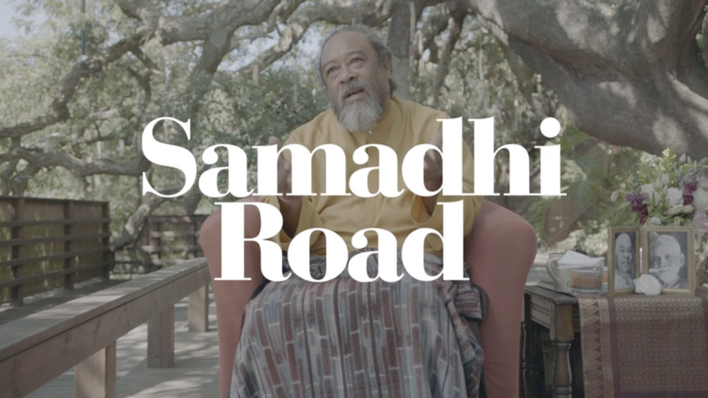 Foto de um senhor de barba branca longa sentado em uma cadeira. à frente, o nome Samadhi Road está nas cores brancas. 