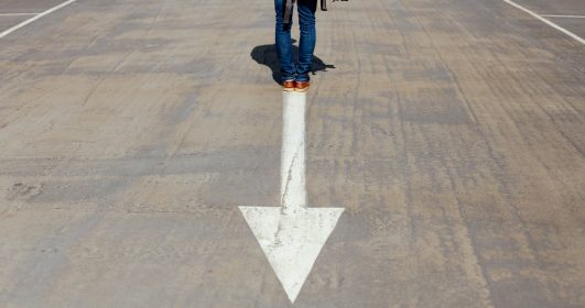 Foto de uma pessoa parada em uma seta orientada para o sul em uma estrada. Há apenas as pernas a mostra e uma mochila nas costas. Vida com propósito
