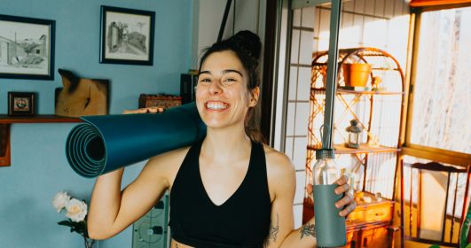 Mulher sorrindo segurando um tapete de prática de yoga e uma garrafa de água antes de se exercitar.
