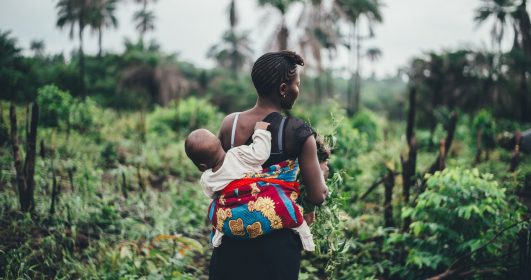 Uma mulher negra carrega seu filho nas costas em uma região de floresta. A agroecologia pode nos salvar da crise climática.