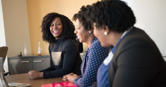 Mulheres negras sorriem juntas em uma mesa de trabalho. O que você pensa sobre networking?