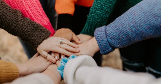 Foto de várias mãos unidas juntas. Cada pessoa veste uma espécie de casaco, embora a imagem só mostre os braços, em alusão ao Dia Internacional da Juventude.