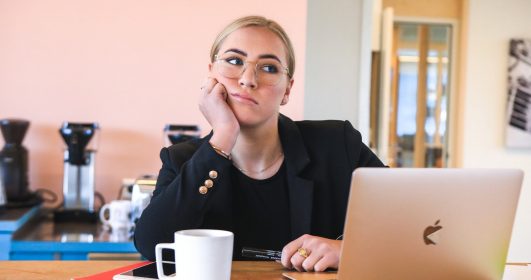 Uma mulher de pele branca, cabelo preso loiro e usando óculos está em frente a um laptop com cara de pensamento. Pensamentos, máximas e provérbios.