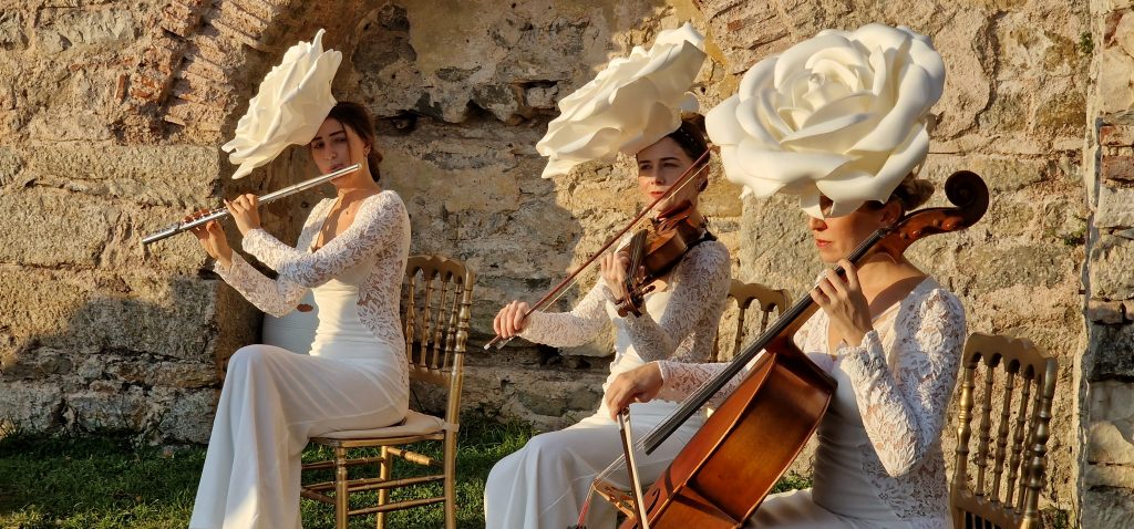 Mulherusam chápeus em formato de flores enquanto estão sentadas em cadeiras tocando violino.