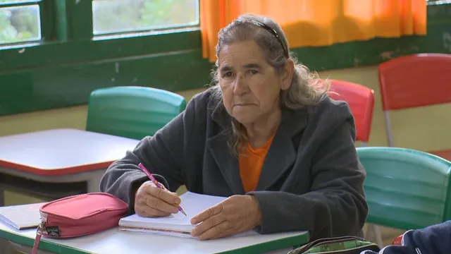 Sônia Teresinha está sentada em uma cadeira da escola com as mãos sobre o caderno, segurando um lápis na mão direita, vestindo um casaco cinza e uma tiara preta no cabelo grisalho. aprender