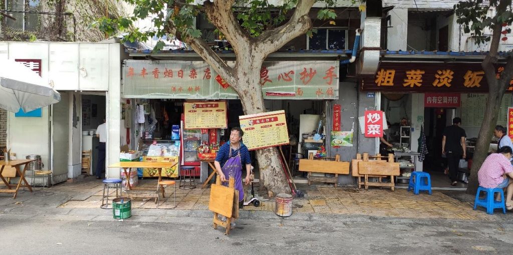 Ruas da cidade de Chengdu, China. Há bancos, lojas, um homem parado na rua, uma árvore e letreiros escritos em mandarim