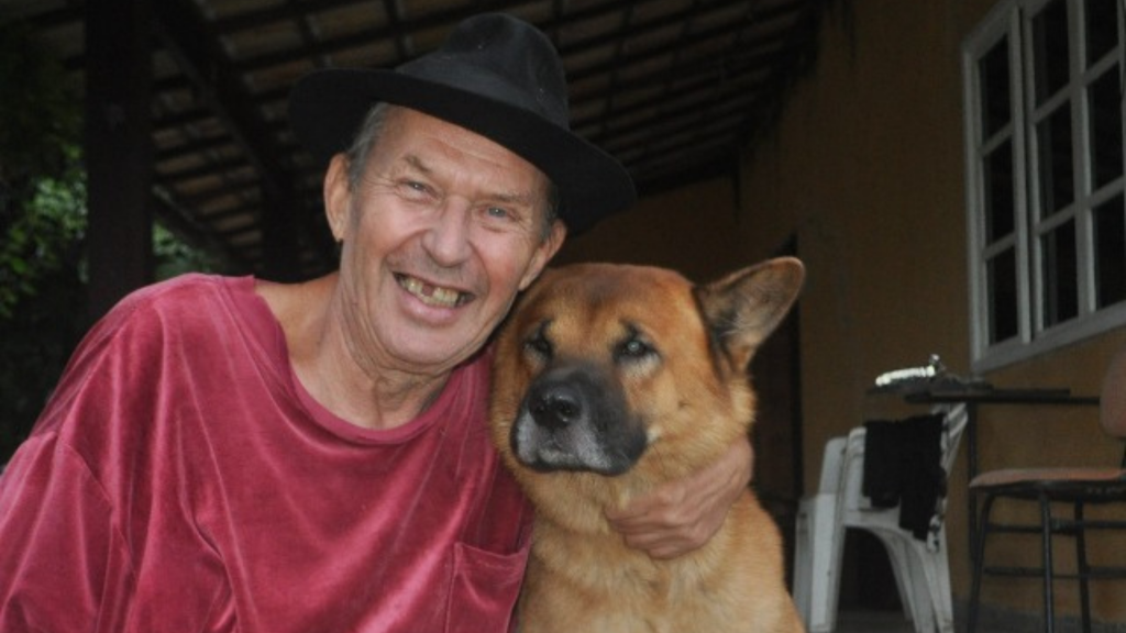 William usa um chapéu, está sorrindo e abraçado com um cachorro de pelo marrom. William também usa uma camisa vermelha. Brasil