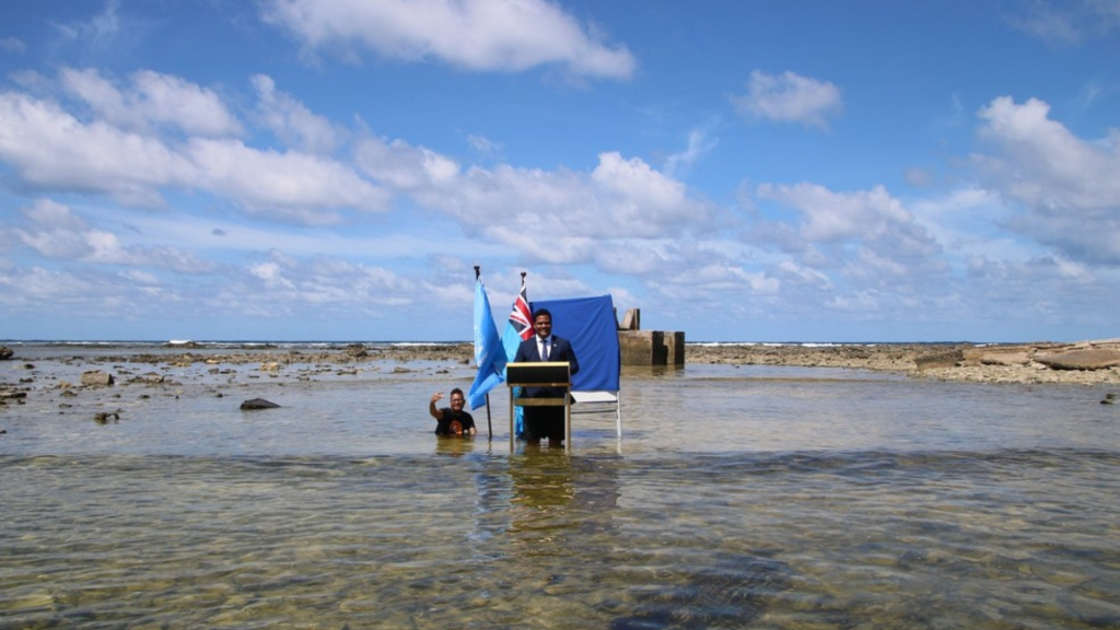 Simon Kofe está no meio do mar enquanto posa para a foto usando um terno, um pulpito e a bandeia do país ao fundo. ruptura climática