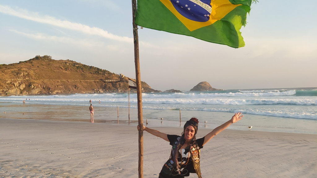 Uma mulher está sorrindo em uma praia com a bandeira do Brasil tremulando. motochileira