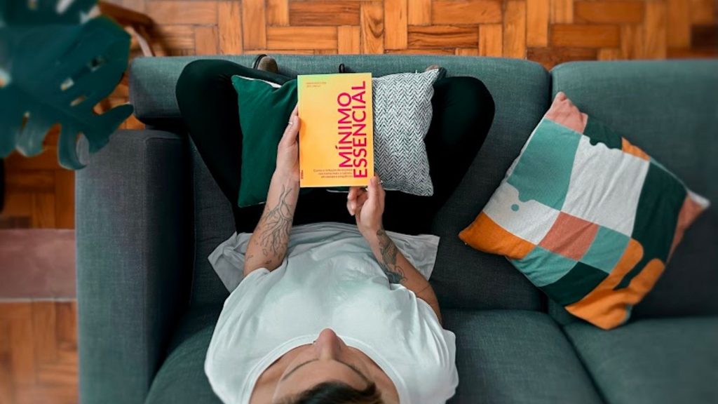 um homem está deitado em um sofá e segura o livro mínimo essencial, em letras vermelhas com capa amarela