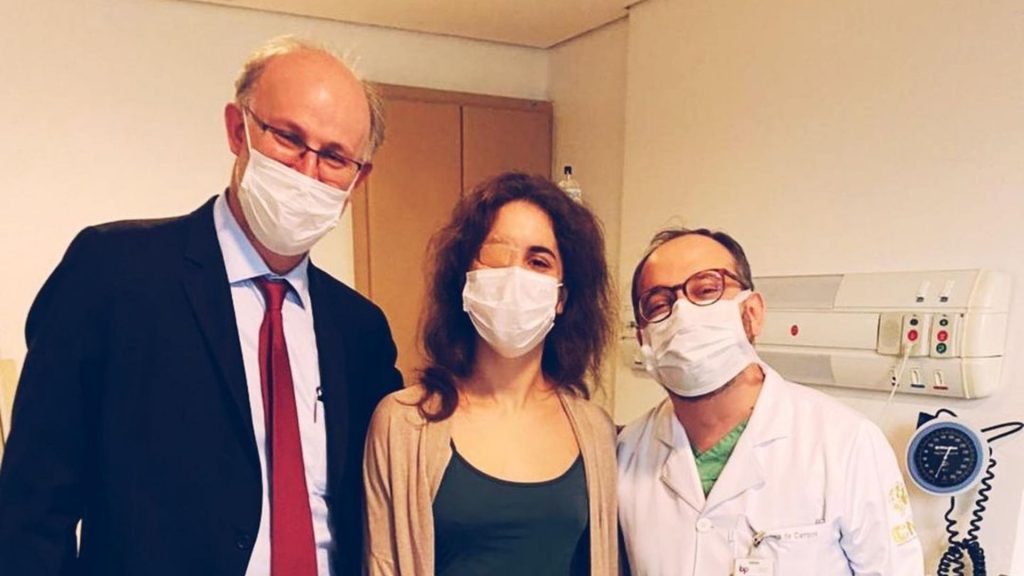 uma mulher, um médico e um homem de palitó preto em uma sala de hospital. Todos usam máscaras. vida