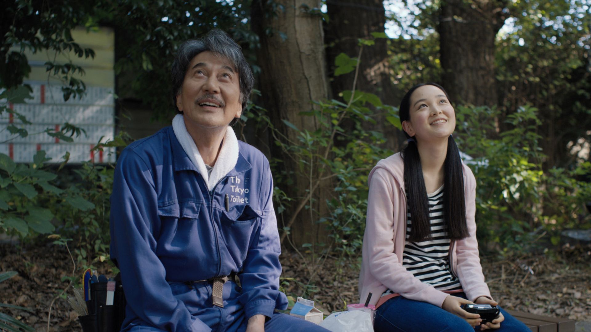 A imagem mostra um homem e uma jovem sentados em um ambiente ao ar livre, cercados por árvores e folhagens. O homem veste um macacão azul com um bordado que diz "The Tokyo Toilet" e aparenta estar sorrindo, olhando para cima. A jovem ao seu lado, também sorridente, está vestida com um casaco rosa claro e uma blusa listrada preta e branca. Ela segura um controle de videogame. Ambos estão sentados lado a lado, em um cenário que transmite tranquilidade e leveza.