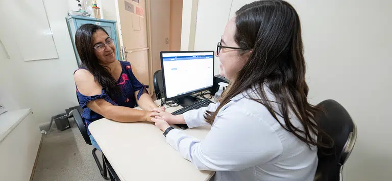 A imagem mostra duas pessoas sentadas em lados opostos de uma mesa de escritório, onde há um computador. À esquerda, uma paciente sorri e parece tranquila, vestindo uma blusa azul com detalhes em rosa. À direita, uma profissional médico de família, usa jaleco branco e óculos, segura as mãos da paciente de maneira carinhosa, transmitindo uma sensação de acolhimento. Elas estão em uma consulta médica em um ambiente claro e organizado, sugerindo um atendimento humanizado.