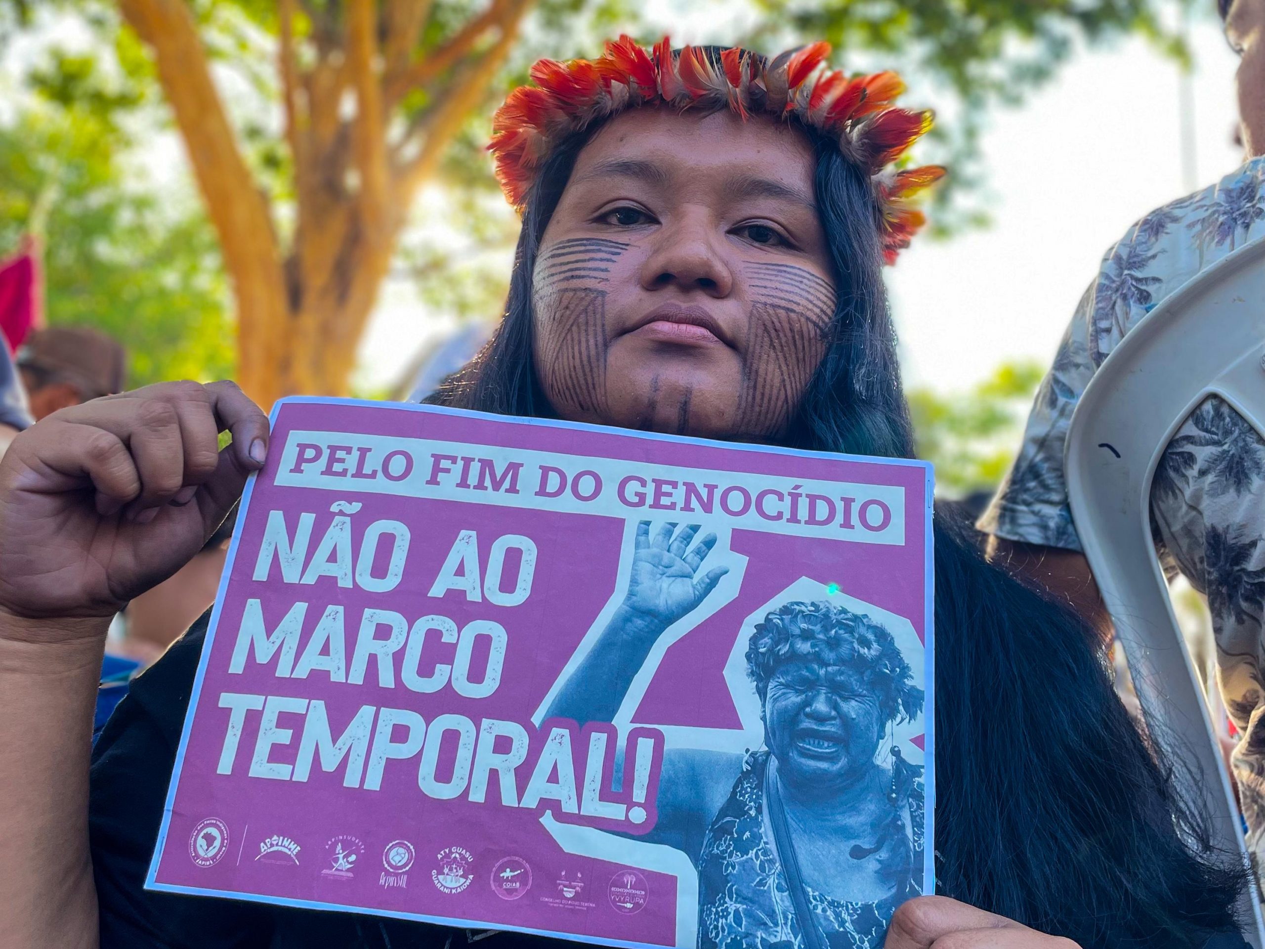 De cocar e pintura facial, a professora indigena Glycya segura um cartaz em protesto contra o marco temporal, onde se lê "Pelo fim do genocídio - não ao marco temporal!"