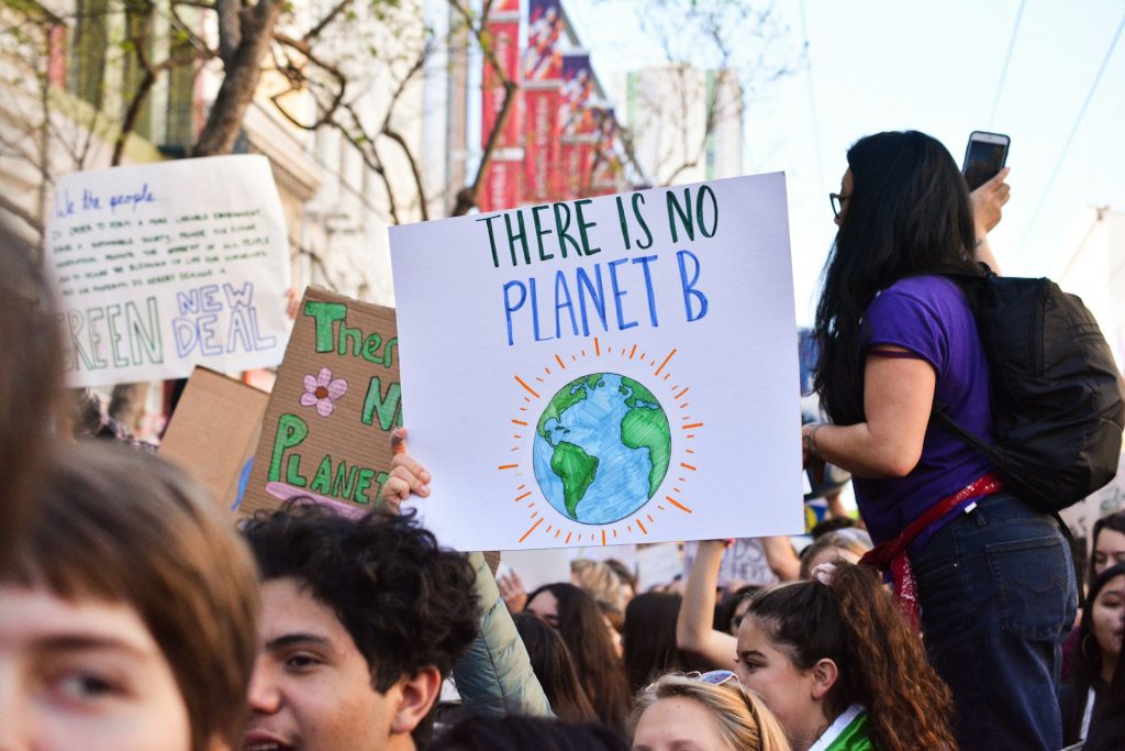 Uma multidão de jovens em uma manifestação. Em destaque, uma pessoa segura um cartaz com a frase em inglês "There is no Planet B" (Não há um Planeta B), acompanhada de uma ilustração do planeta Terra. Ao fundo, outros cartazes fazem referência à crise climática e à necessidade de ações sustentáveis para proteger o meio ambiente.