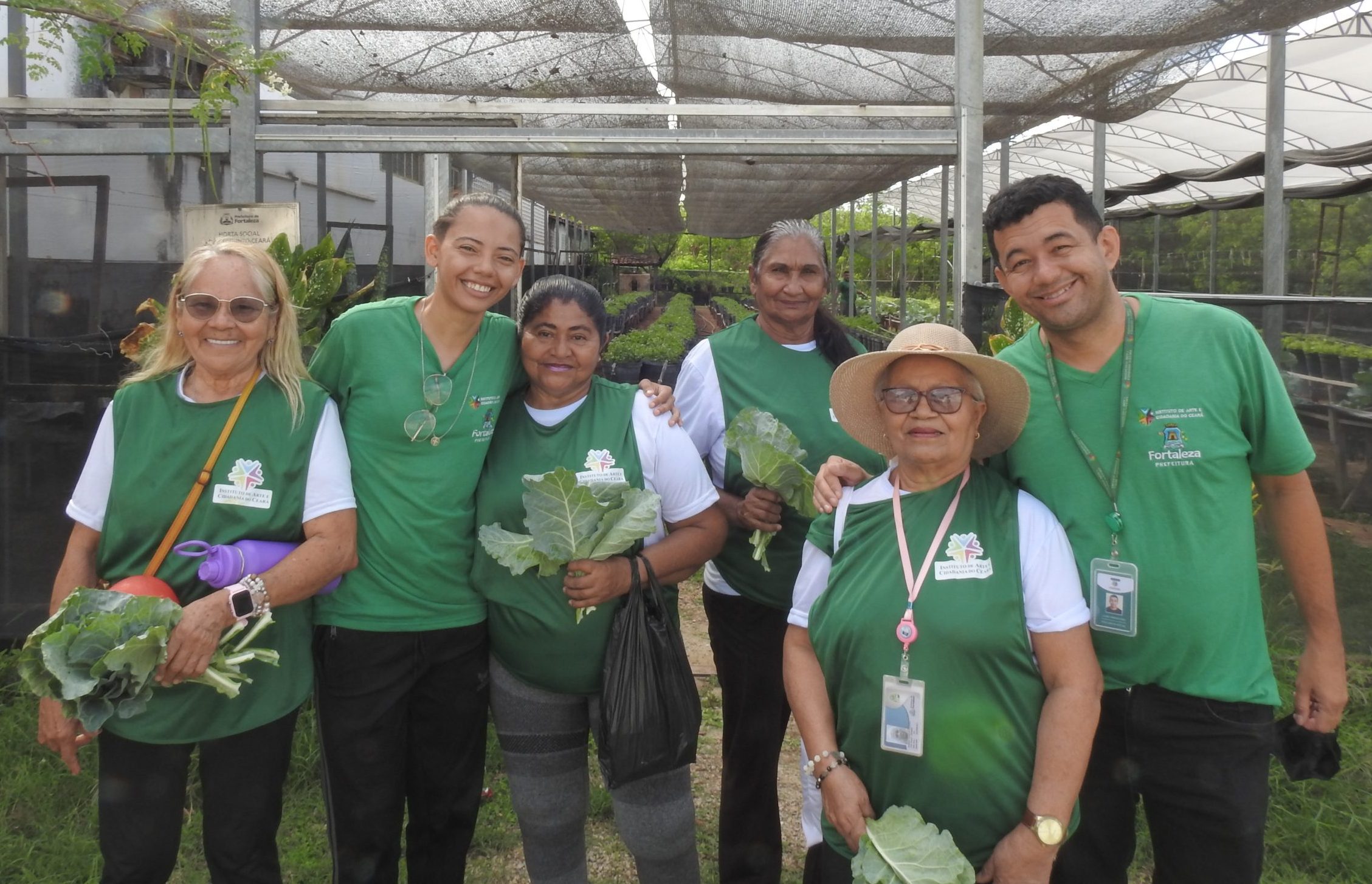 Uma foto de seis pessoas sorridentes em uma horta comunitária. Todas estão vestindo camisetas verdes com o logo de um projeto agrícola local, em Fortaleza, conforme as identificações. O grupo é composto por quatro mulheres e dois homens, de idades variadas. Uma das mulheres usa chapéu e óculos escuros, e segura folhas de couve. Outra senhora segura um molho grande de couve e uma garrafa de água roxa. Todos parecem felizes e descontraídos, abraçados e posando juntos. Ao fundo, há estufas cobertas com telas para cultivo de plantas, em um espaço organizado e verde.