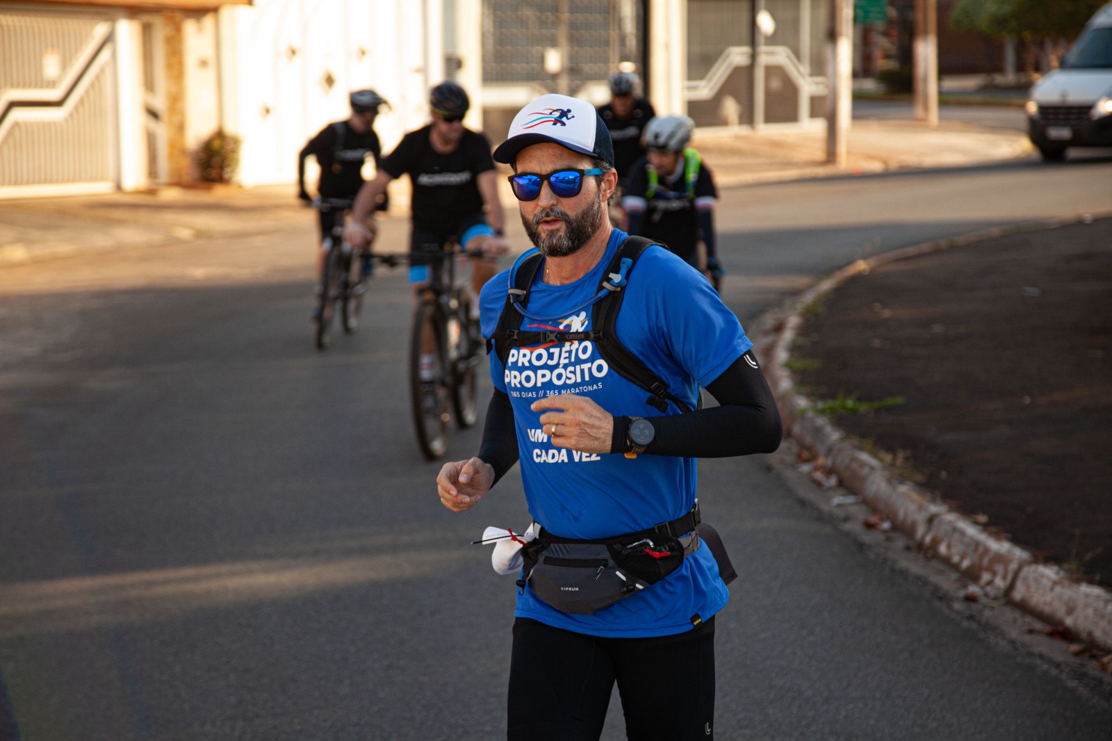 Na imagem, há um homem a correr em uma rua asfaltada durante o dia. Ele está vestindo uma camiseta azul com a frase "Projeto Propósito" estampada no peito, além de um boné branco com detalhes azuis e óculos escuros espelhados. Ele também usa um colete com suporte de hidratação e uma pochete na cintura. Atrás dele, algumas pessoas estão pedalando bicicletas, vestidas em roupas esportivas. A cena ocorre em um ambiente urbano com árvores, calçadas e uma van estacionada ao fundo.