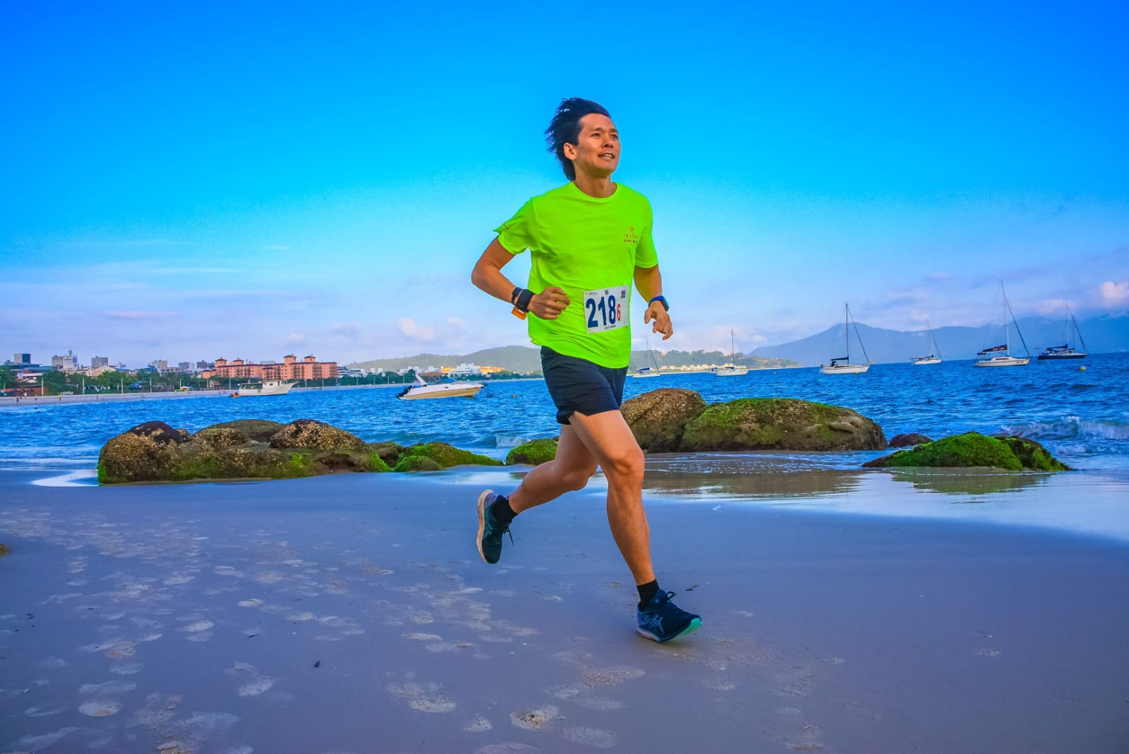 Foto de uma pessoa correndo na areia de uma praia durante o dia. A pessoa veste uma camiseta verde neon com o número 218 preso na frente, shorts preto e tênis de corrida escuros. Ao fundo, há um mar azul com algumas pedras cobertas de musgo e vários barcos ancorados. O céu está limpo e azul, com montanhas ao longe, sugerindo um clima agradável e ensolarado. A pessoa tem