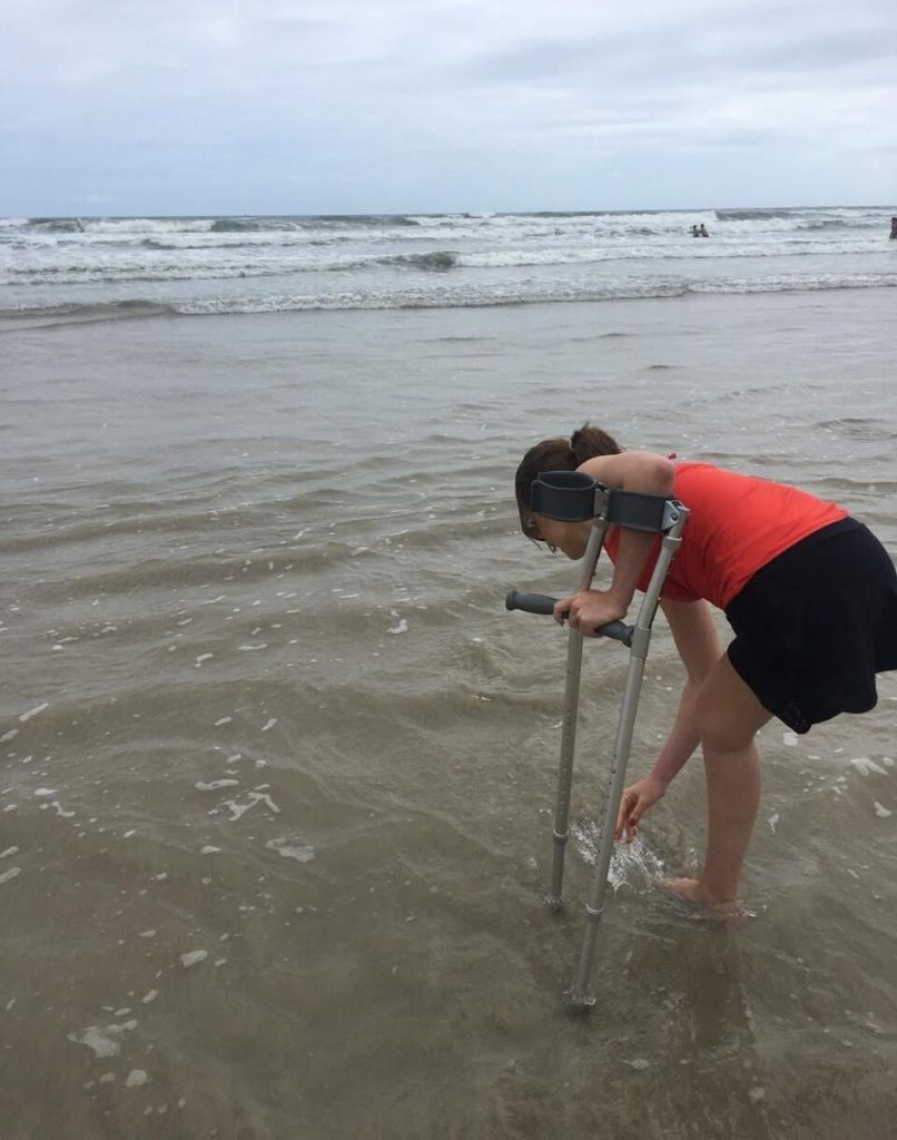 Gabriela apoiada na muleta e com o pé na água do mar, abaixada para molhar a mão