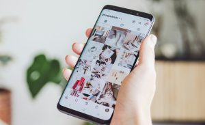 Pessoa segura um smartphone exibindo um perfil do Instagram com imagens organizadas e esteticamente agradáveis. A cena destaca o papel das redes sociais na curadoria visual e na promoção de estilos de vida.